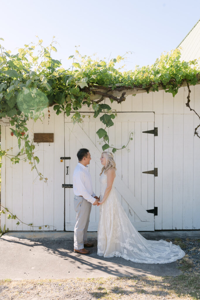 Bride and groom holding hands under grapevines by a white barn at Chateau Lill.