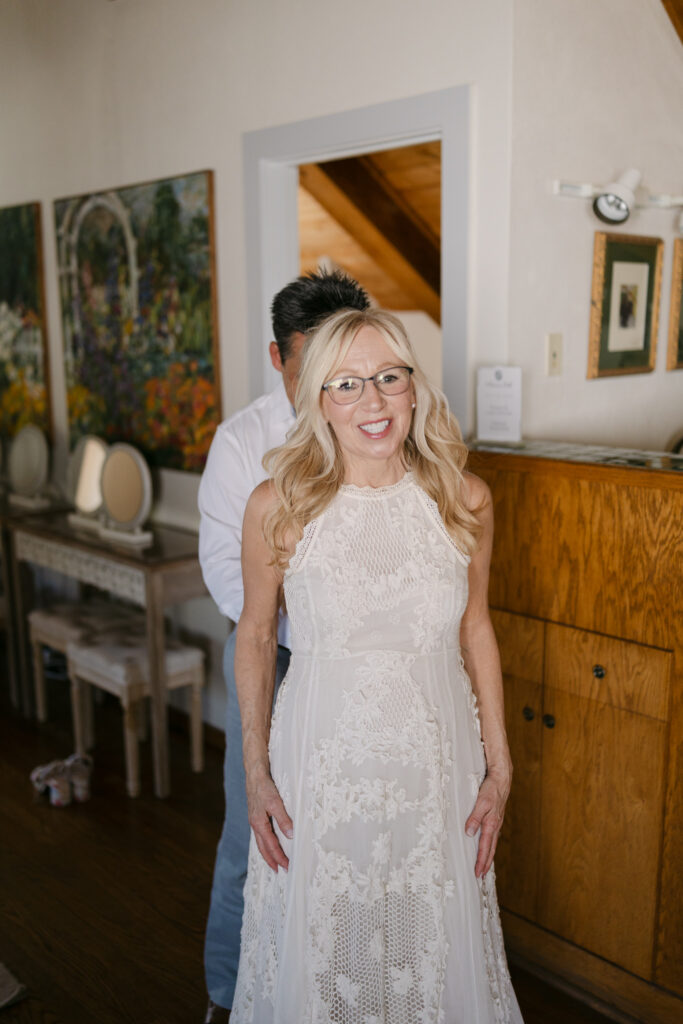 Bride smiling while groom helps button her lace wedding dress at Chateau Lill.