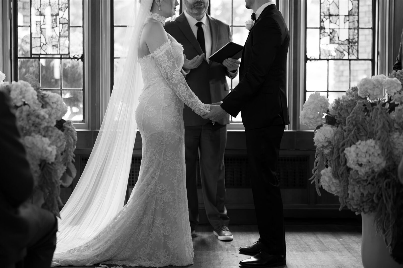 Black and white wide shot of a bride and groom holding hands during their indoor wedding ceremony in the Great Hall at Thornewood Castle wedding venue.