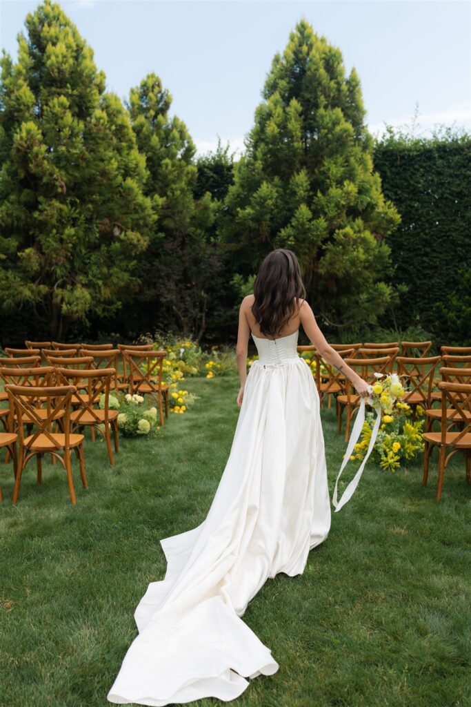 Bride walking through ceremony aisle with wooden chairs and yellow florals at Briarwood Estate wedding.