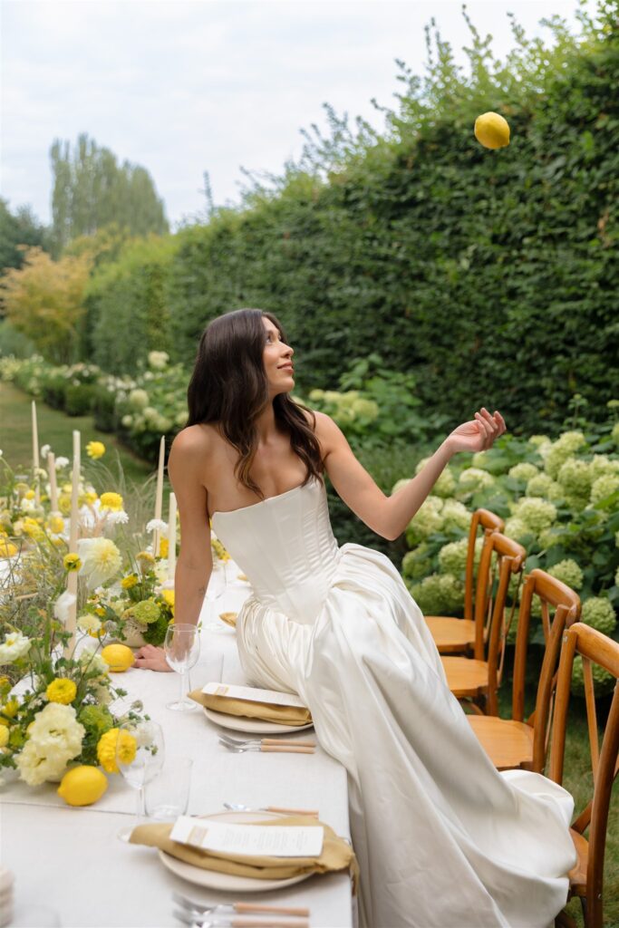 Bride sitting on reception table in garden setting at Briarwood Estate wedding while tossing a lemon in the air.
