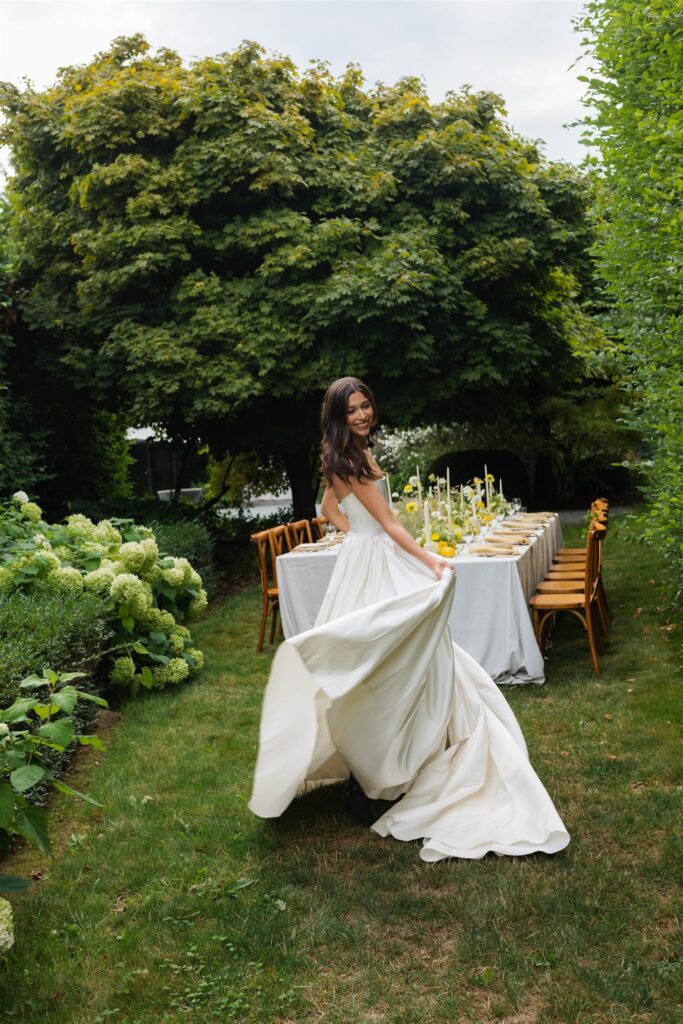Bride posing for portraits in a garden area in front of a styled wedding reception table setup. 
