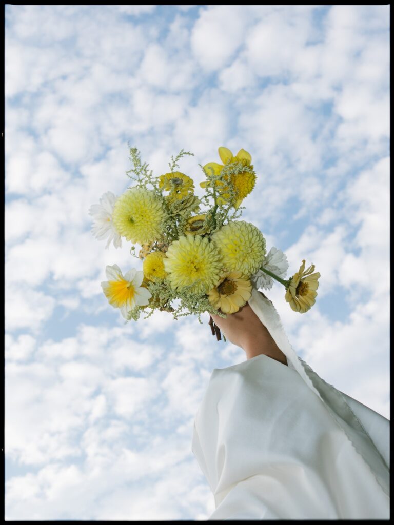 Bride holding her yellow bouquet up in the air with views of the sky.