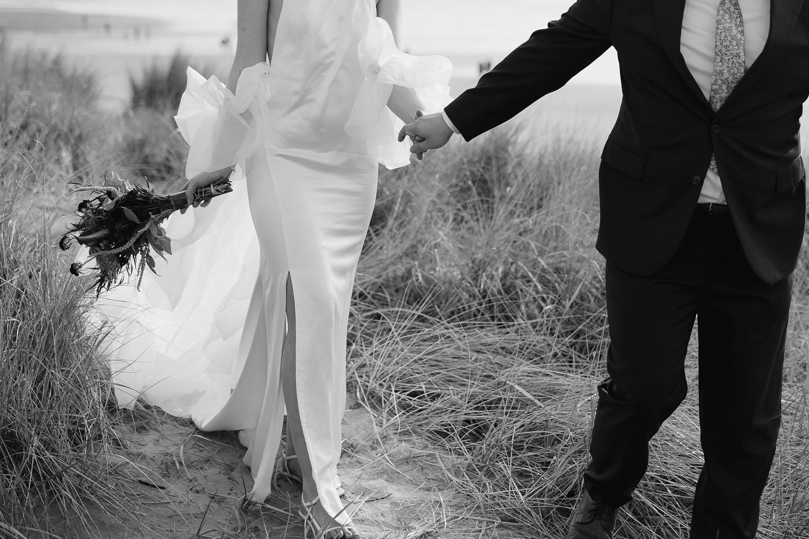 Black and white wide shot of a bride and groom holding hands and walking at Cannon Beach for their elopement.
