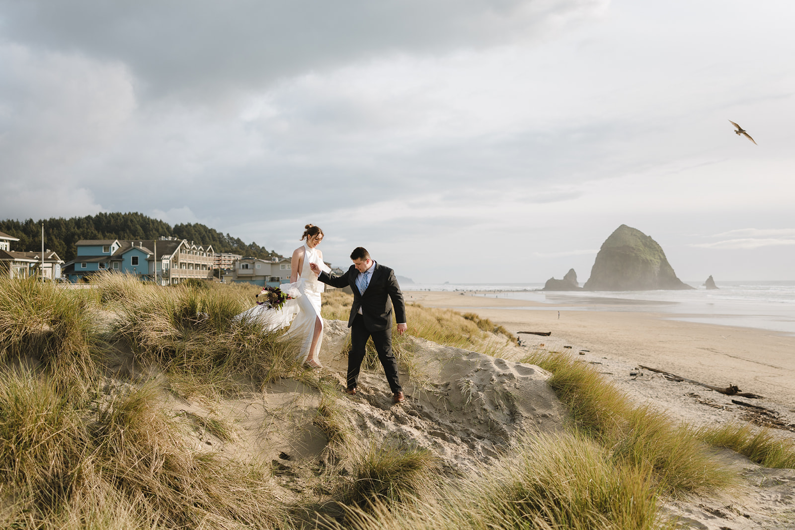 Groom helping the bride walk down a sand dunes at Cannon Beach with houses and Haystack Rock in the distance. 