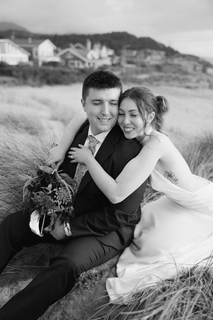 Black and white portrait of a bride and groom sitting in the beach grass.