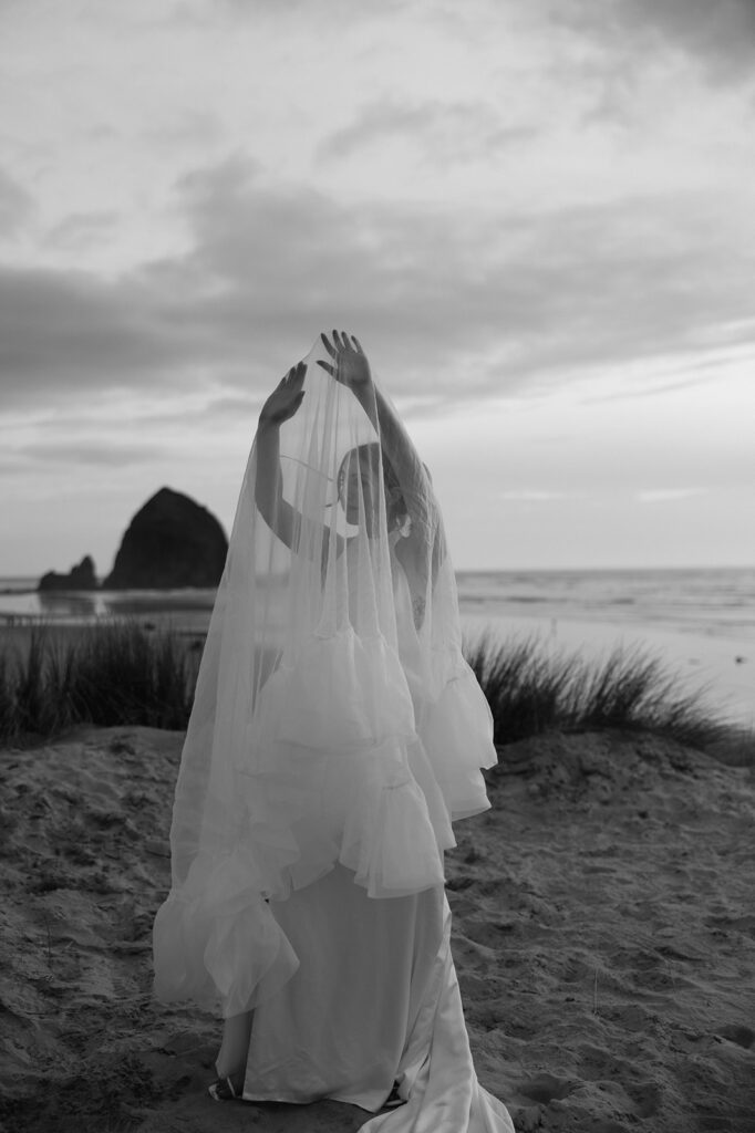 Black and white portrait of a bride holding up her veil over her for editorial bridal portraits on Cannon Beach with Haystack Rock behind her in the distance.
