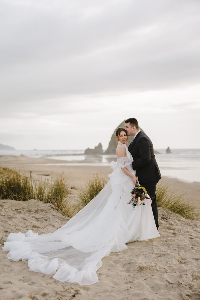 Groom kissing the brides forehead during their Cannon Beach elopement phots with Haystack Rock behind them.