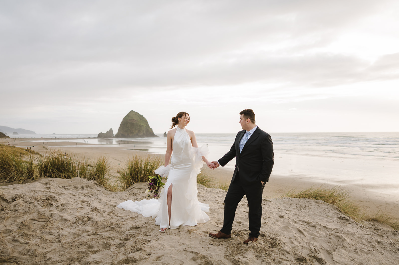 Bride and groom smiling at each other as they hold hands on Cannon Beach with Haystack Rock in the distance.
