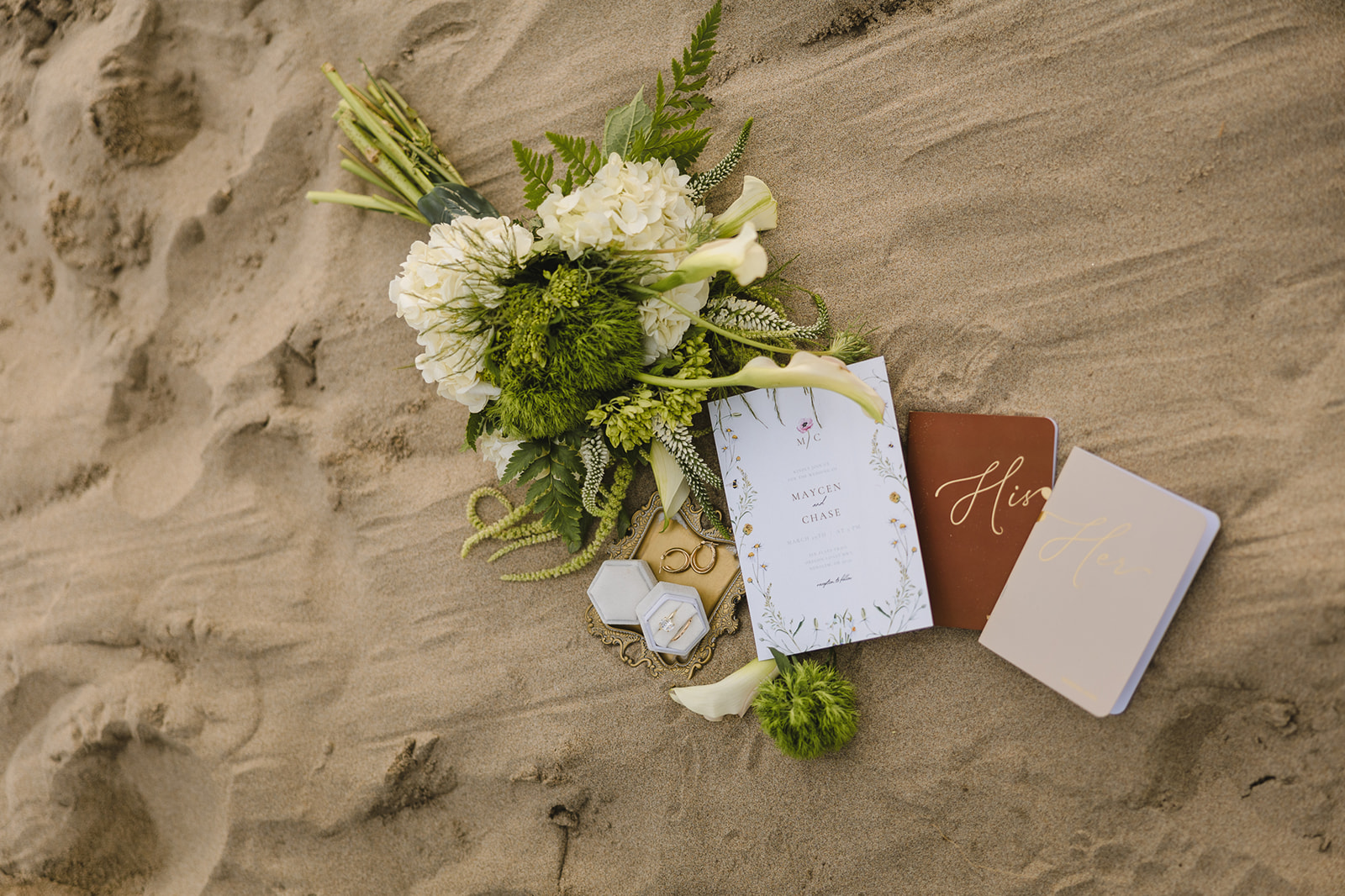Cannon Beach elopement details with invite, vow books, florals. and jewelry styled in the sand.