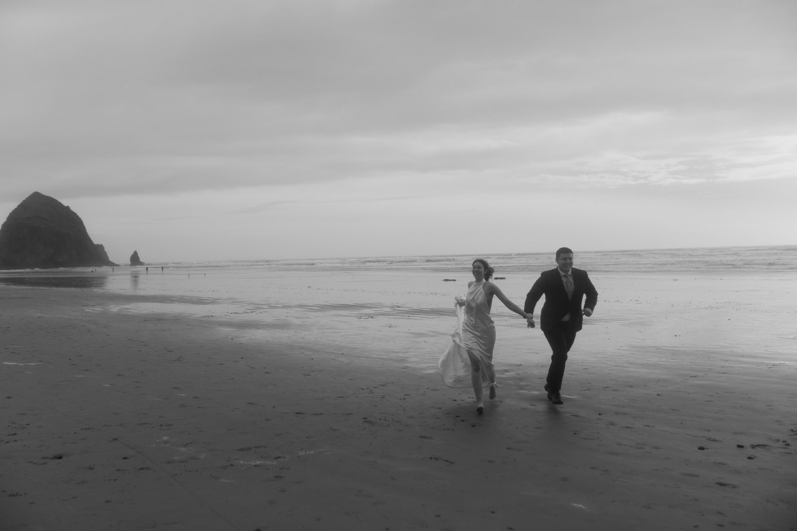 Black and white photo of a bride holding hands and running along the beach with Haystack Rock in the background. 