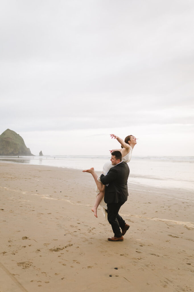 Groom playfully lifting the bride on the beach with Haystack Rock in the distance.