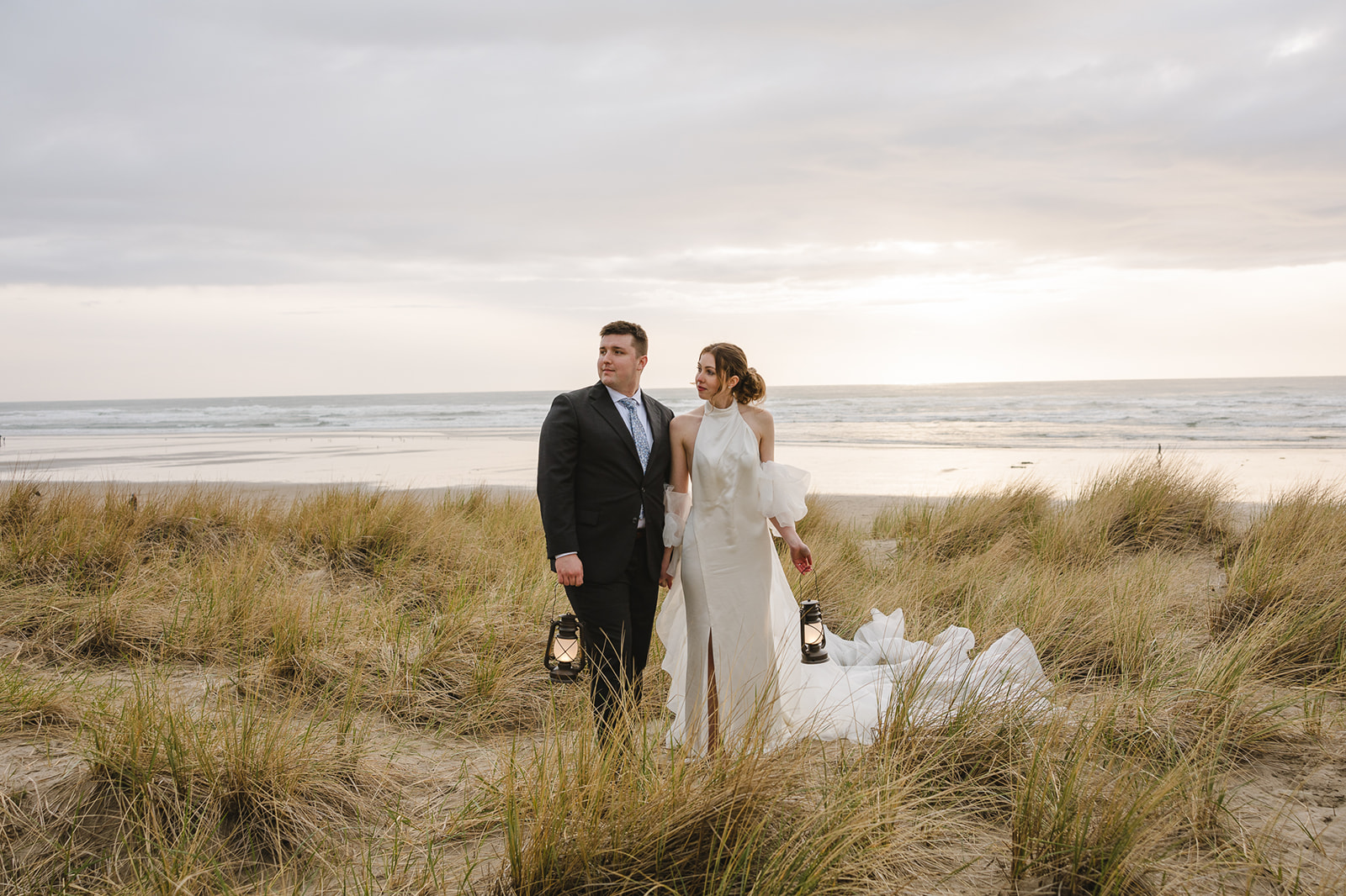Bride and groom standing in the beach grass with the ocean behind them holding lanterns for their Cannon Beach elopement photos.