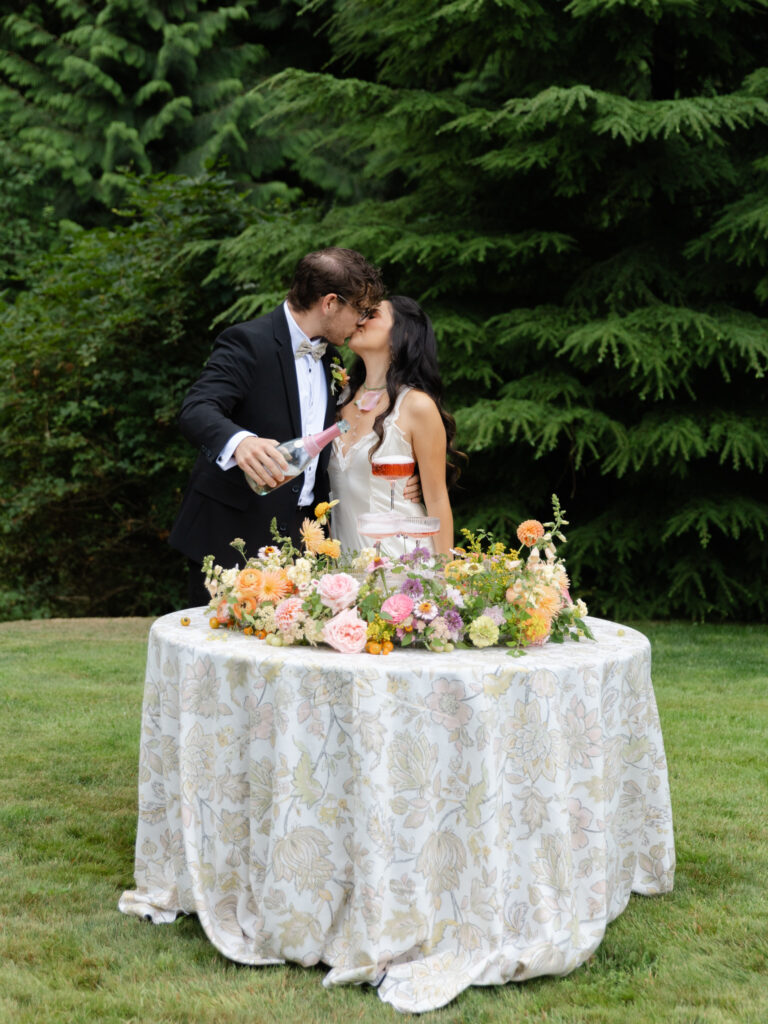 Couple kissing while pouring a champagne tower with colorful flowers at Foxglove Lodge Vashon Island.