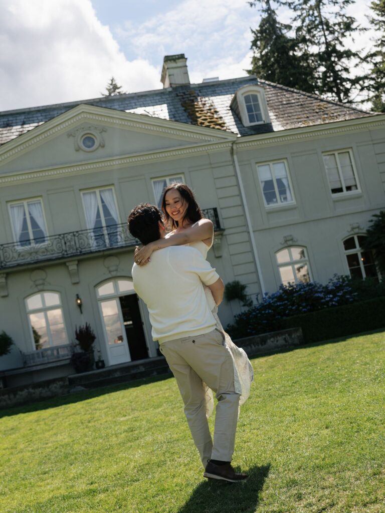 Man lifting his fiancé at Bloedel Reserve with The Residence in the background.