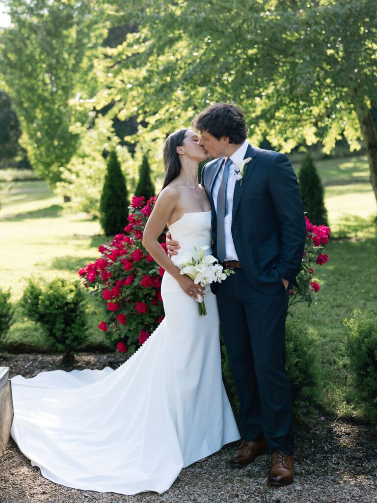 Bride and groom kissing in front of a flower bush at Froggsong Gardens.