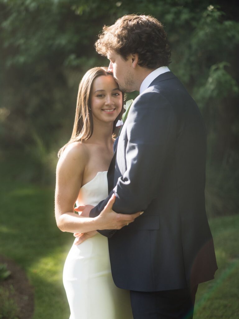 Bride smiling at the camera as the groom kisses her forehead during their golden hour portraits.