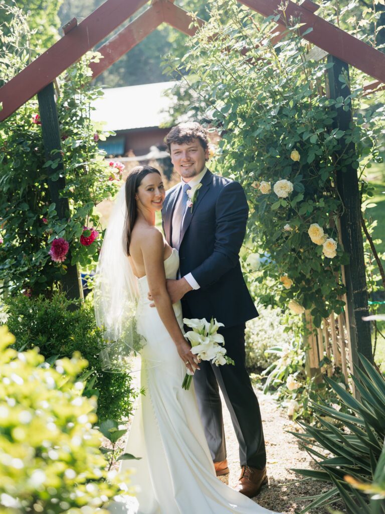 Bride and groom posing beneath a flower-covered garden arch after their ceremony.