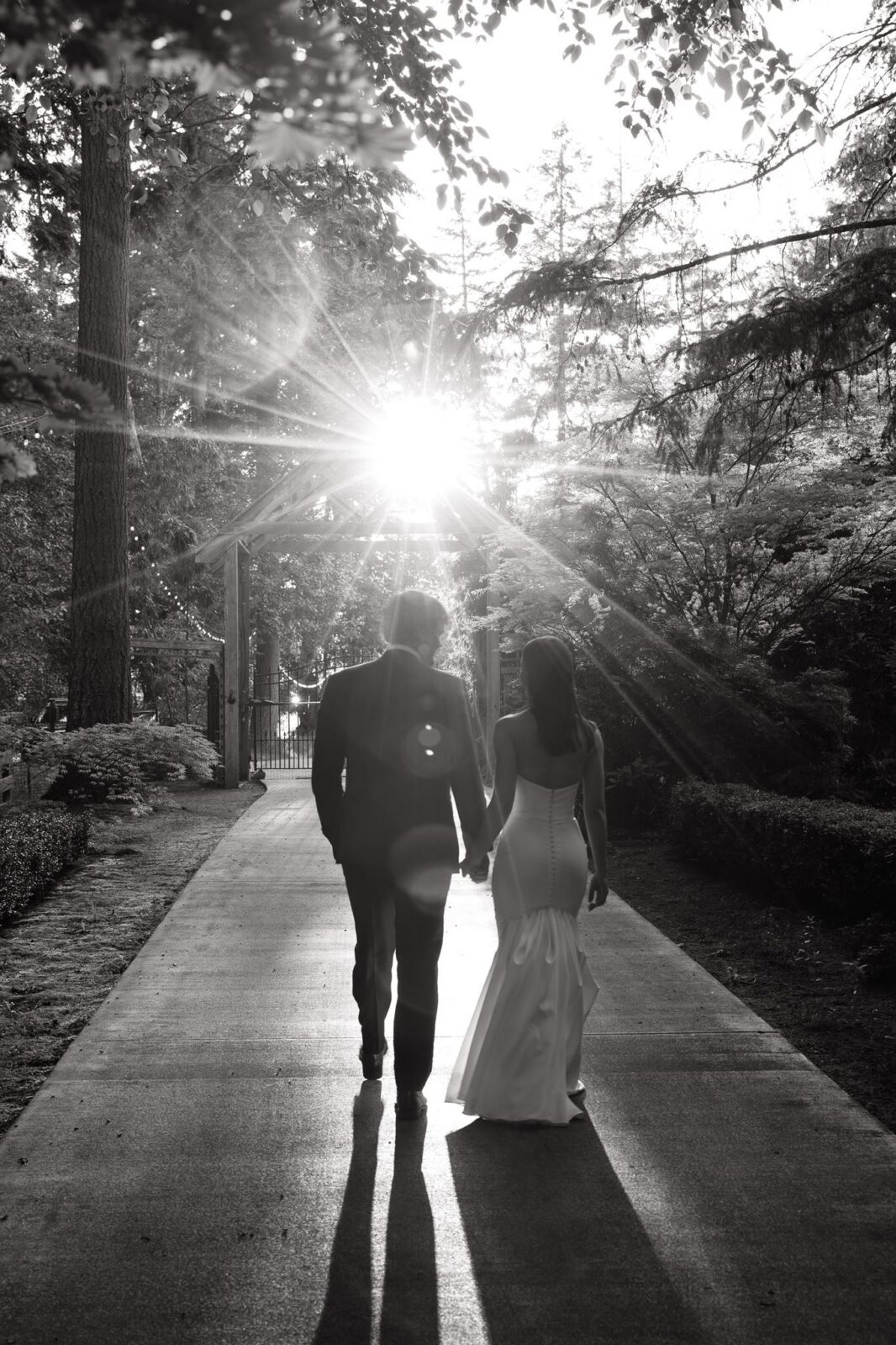 Black-and-white photo of newlyweds walking hand in hand through Froggsong Gardens at sunset.