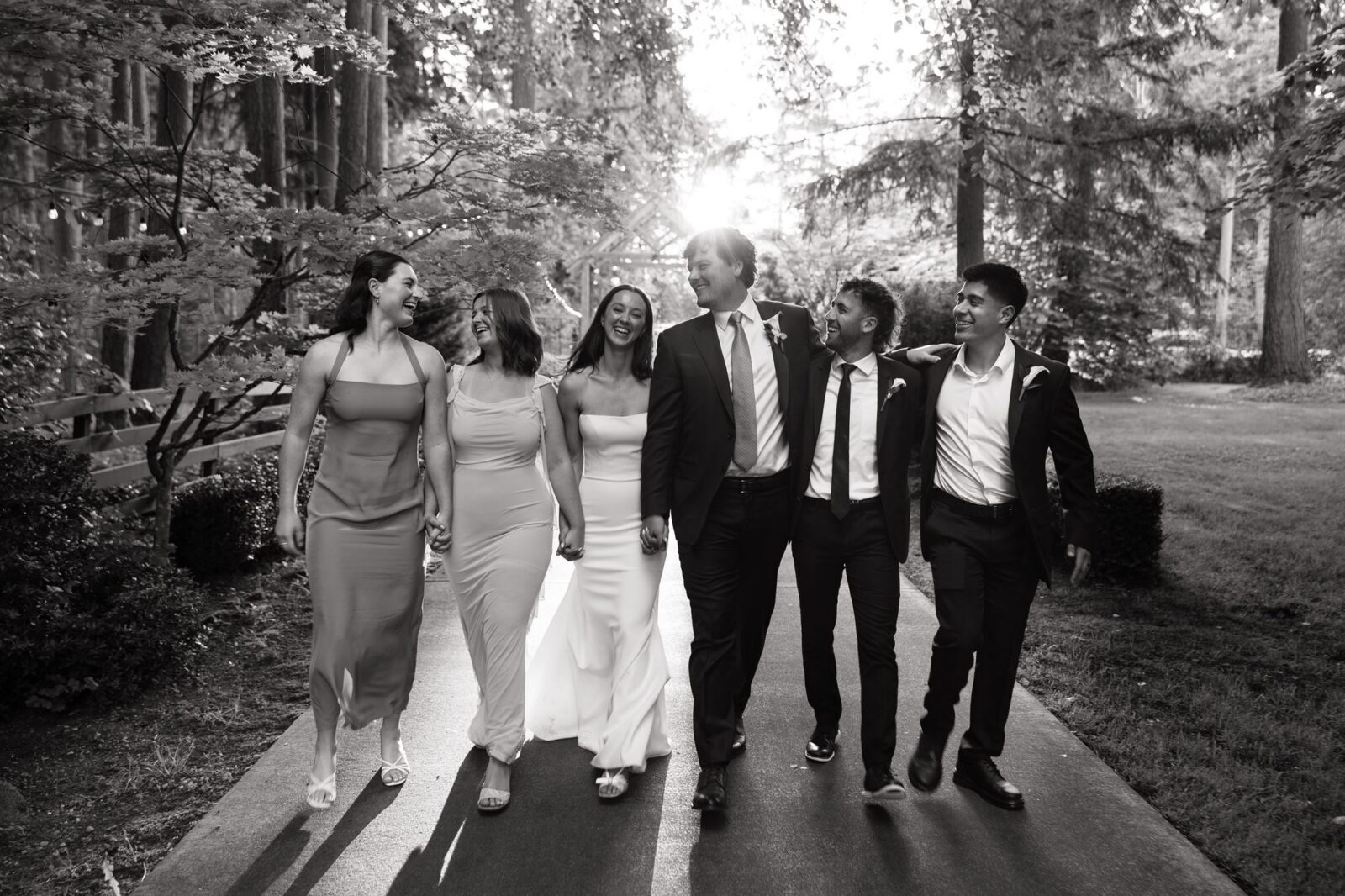 Black and white photo of a bride and groom walking down a pathway with their wedding party at Froggsong Gardens.
