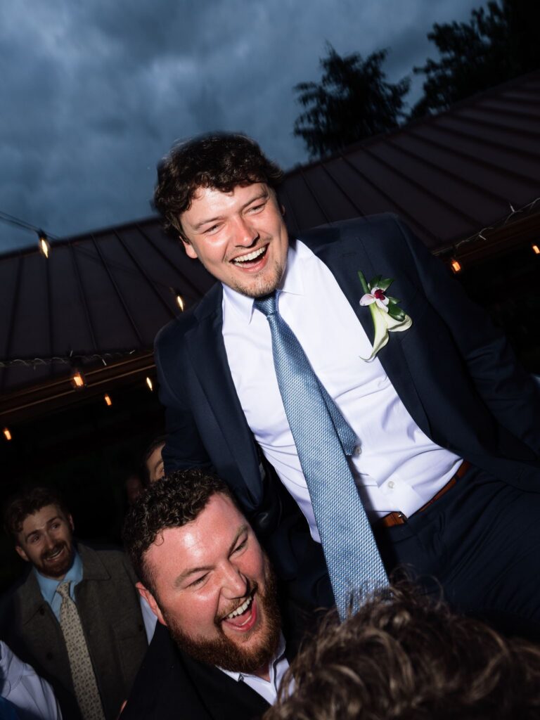 Groom laughing while being lifted onto a guest’s shoulders on the dance floor.