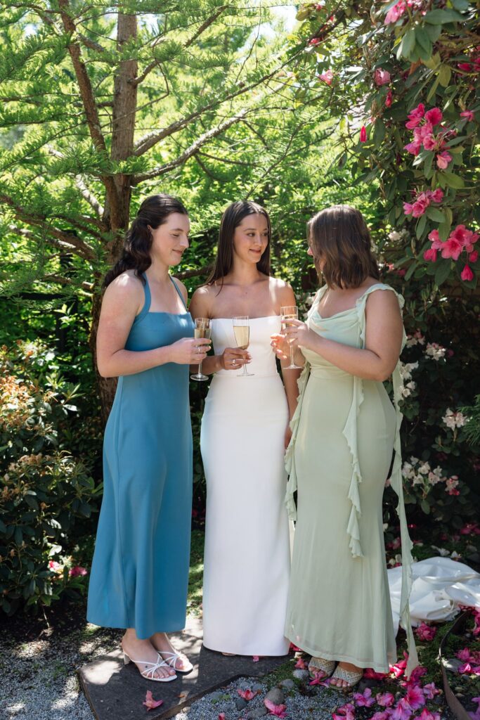 Bride sharing champagne with her bridesmaids beneath pink flowers.