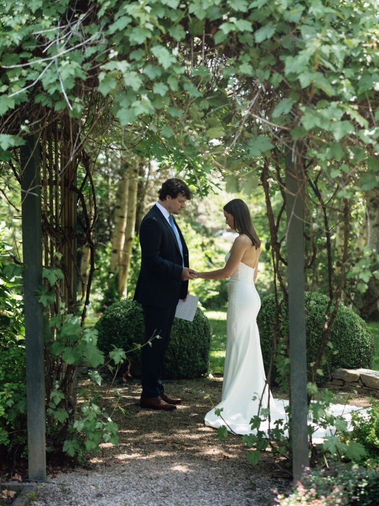 Bride and groom holding hands beneath the greenery at Froggsong Gardens.