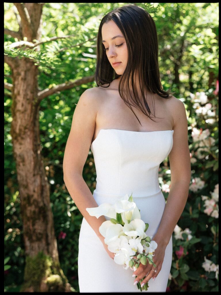 Bride in a strapless wedding dress holding a white floral bouquet in the gardens.