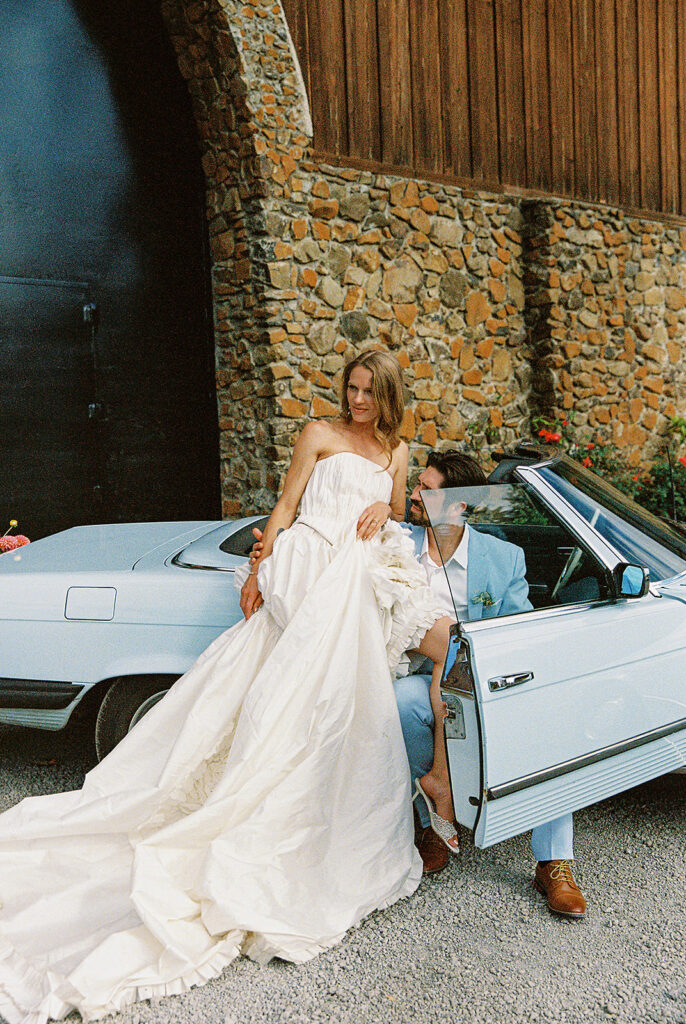 Bride in a textured wedding gown posing beside a groom in a light blue suit and vintage convertible at Maysara Winery.