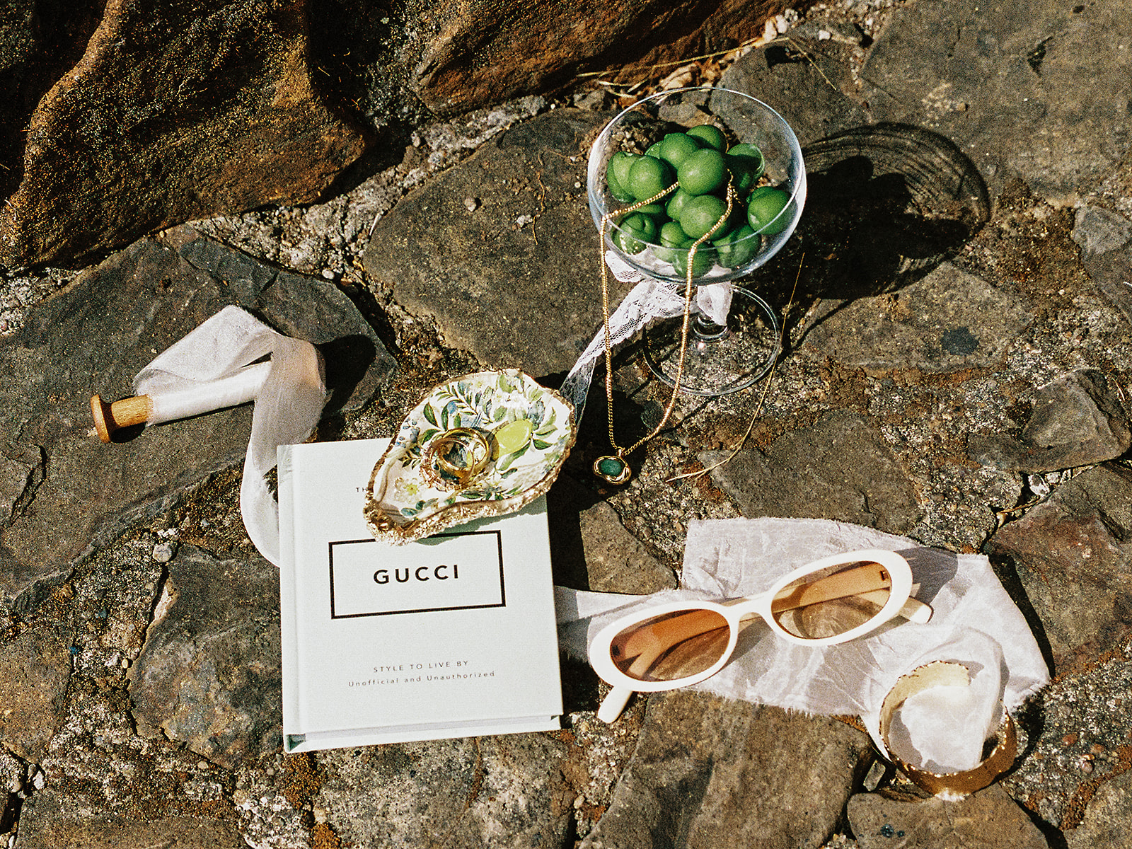 Editorial wedding detail flat lay with sunglasses, jewelry, green grapes, ribbon, and fashion book on stone.
