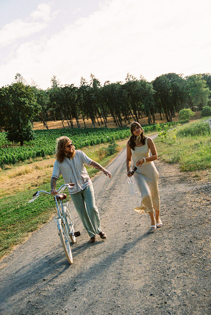 Couple walking side by side with a bicycle along a road surrounded by vineyard rows.