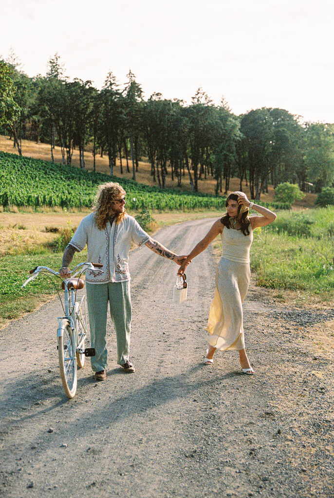Couple walking hand in hand with a bicycle along a road surrounded by vineyard rows.