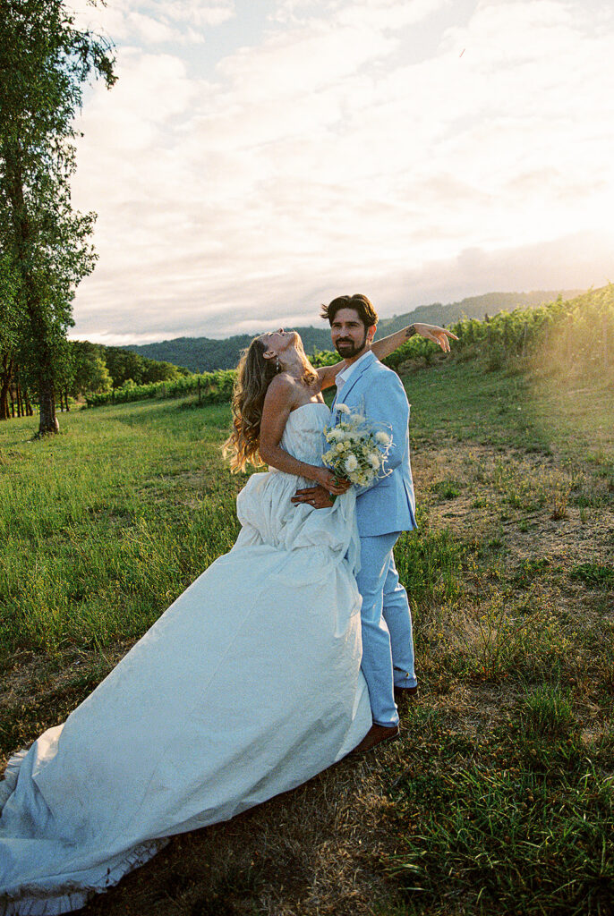 Bride in a textured wedding gown and groom in a light blue suit posing at sunset beside the vineyard.