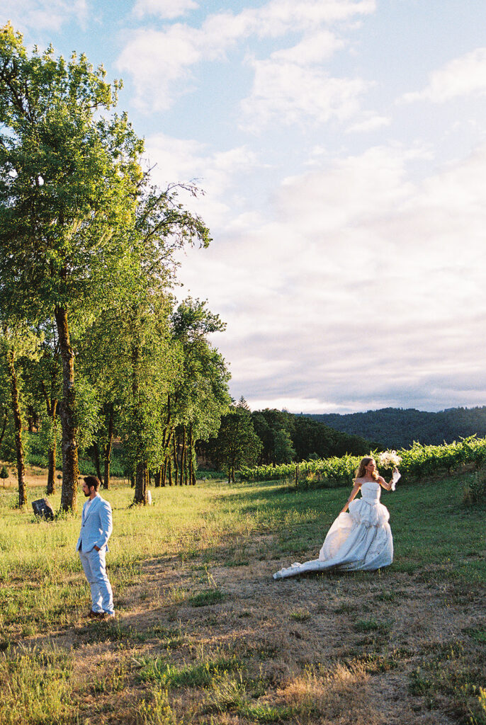 Bride in a textured wedding gown and groom in a light blue suit posing at sunset beside the vineyard.