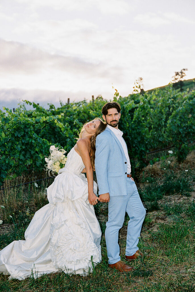 Couple holding hands beside vineyard rows, with the bride in a textured white gown and groom in a light blue suit.
