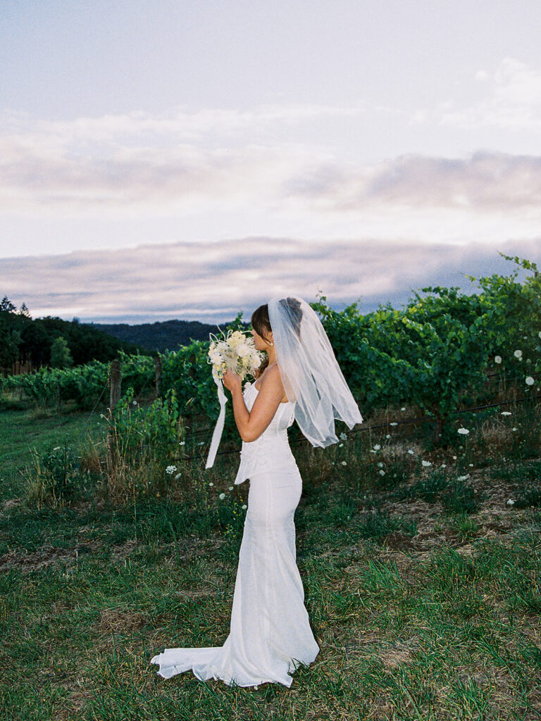 Bride in a textured strapless wedding gown holding a white bouquet in a grassy field at Maysara Winery.