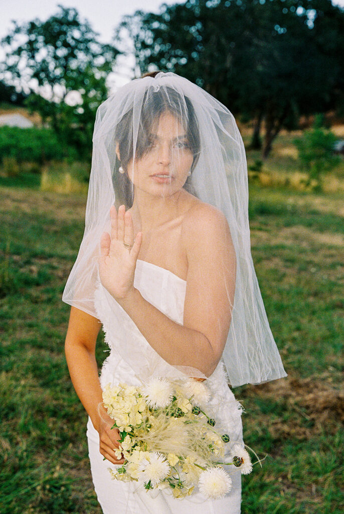 Bride wearing a fitted strapless wedding dress and veil while holding a white bouquet beside the vineyard.